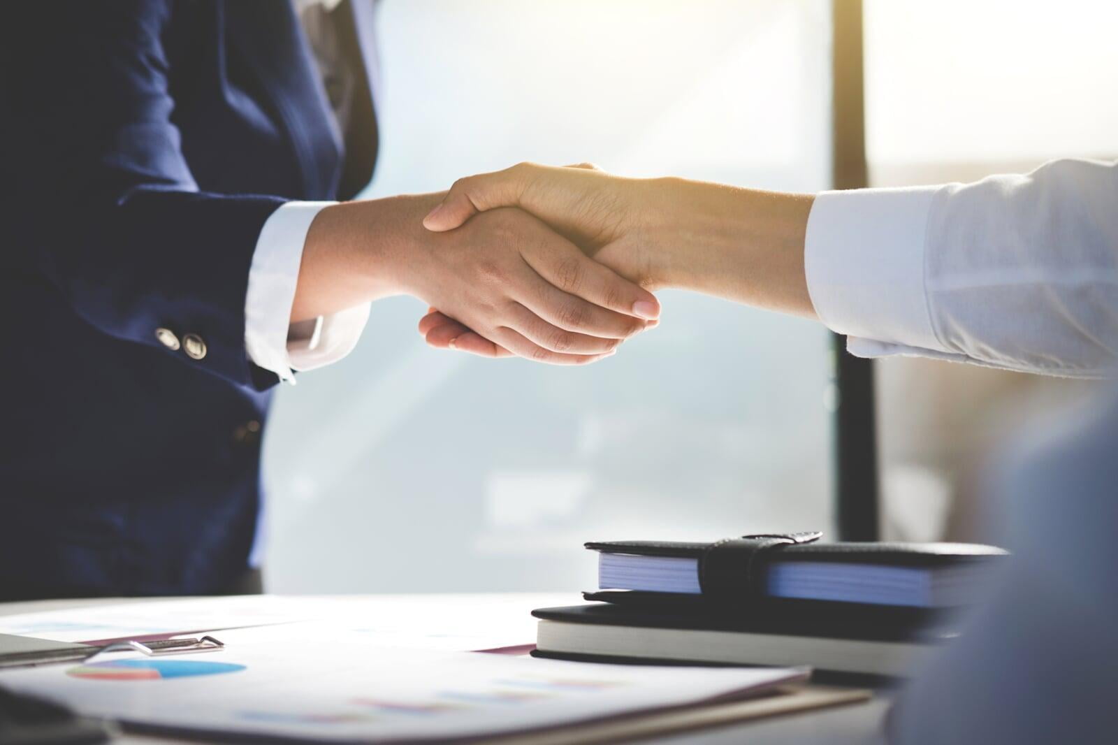 die Arme zweier Frauen beim Handshake, eine hat eine weiße Bluse an, die andere trägt eine weiße Bluse mit einem dunkelblauen Sakko