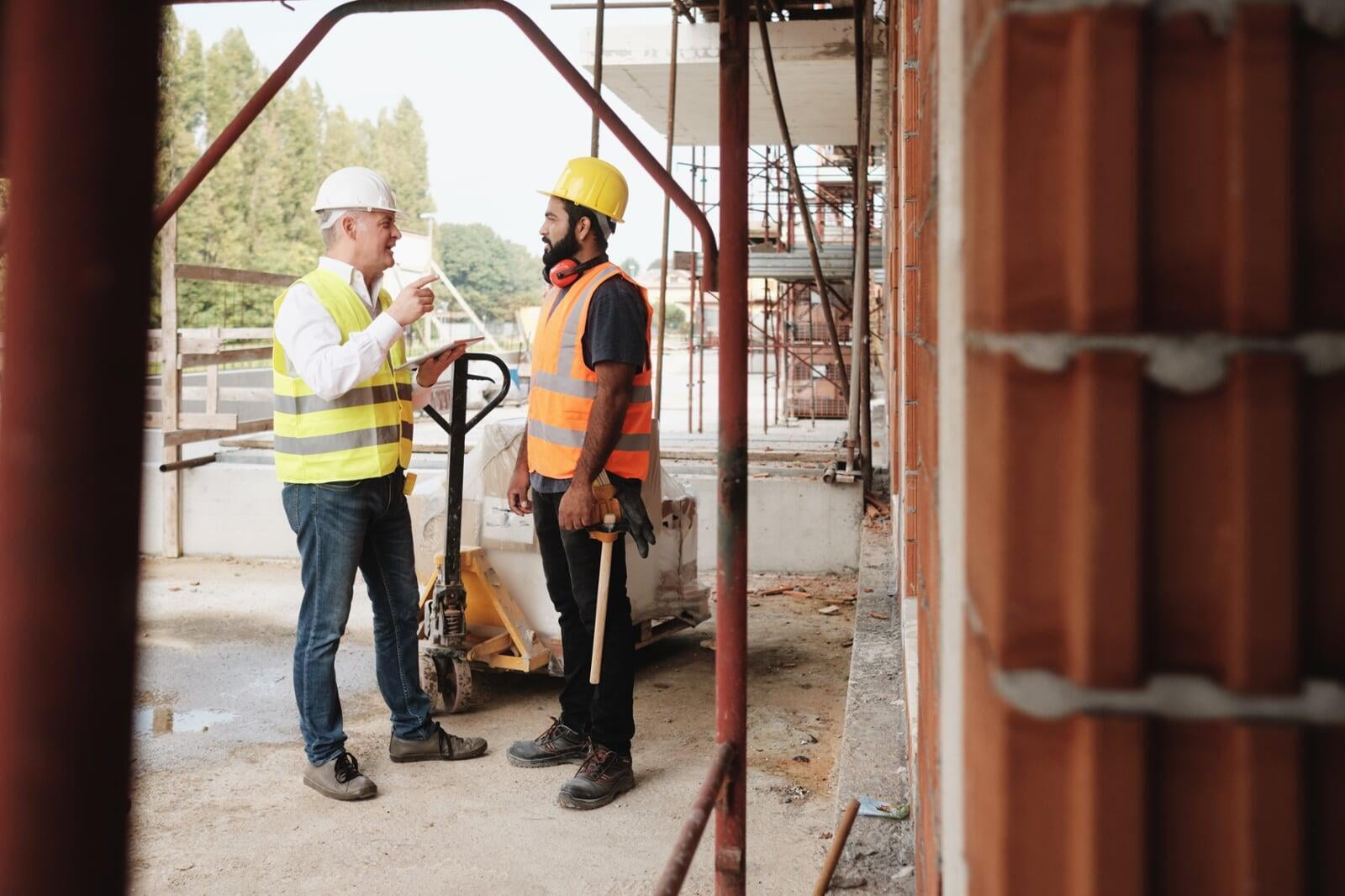 ein Bauingenieur und ein Bauarbeiter mit Bauhelmen beim Gespräch auf einer Baustelle