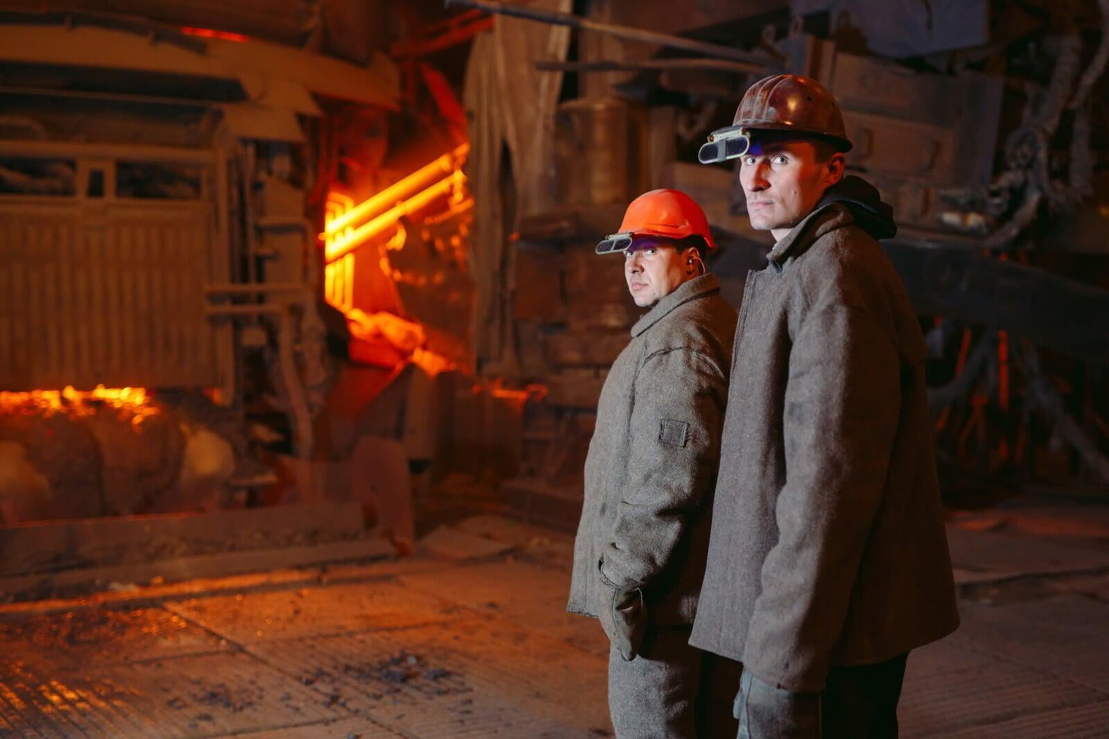 zwei Stahlarbeiter mit Helmen und Schutzkleidung vor Brennofen