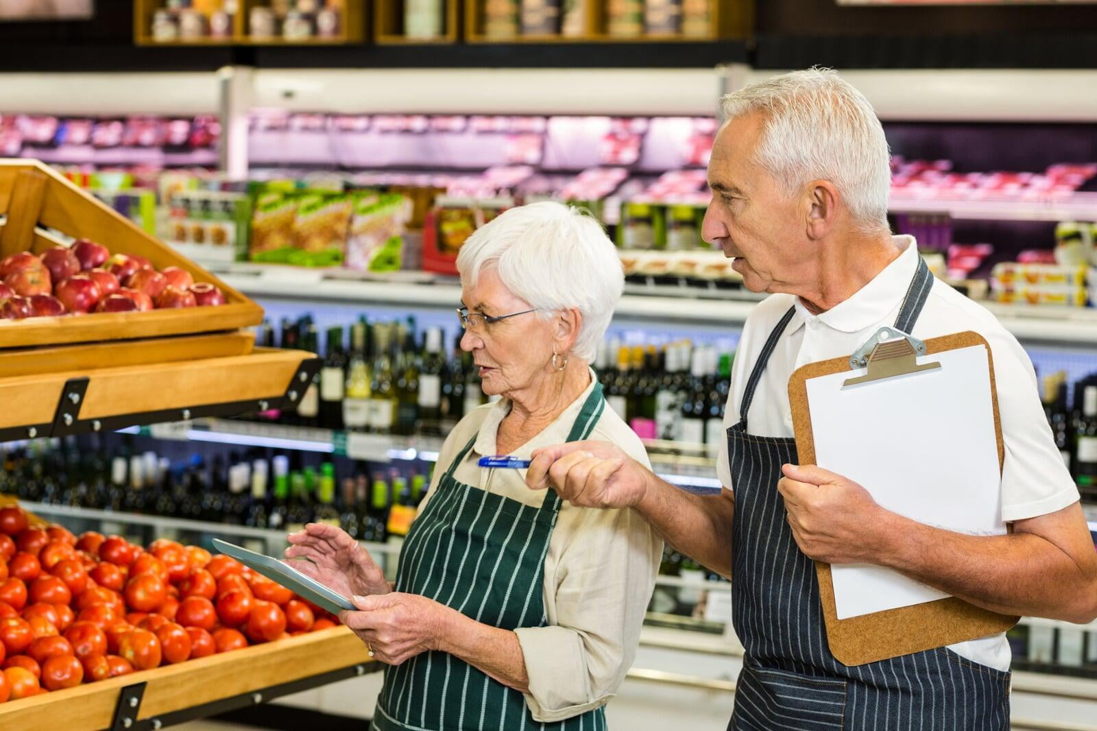 eine ältere Verkäuferin und ein älterer Verkäufer vor einem Obststand in einem Supermarkt